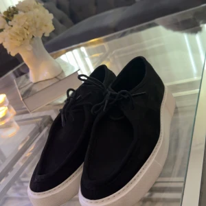 Svarta sneakers med vit sula - Säljer ett par stilrena svarta sneakers i mocka med vit sula. De har snörning och en klassisk design som passar perfekt för både vardag och fest. Skorna är i mycket bra skick och redo för nya äventyr!