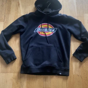 Svart hoodie från Dickies - Säljer en svart hoodie från Dickies med deras klassiska logga i rött och gult på bröstet. Den har en stor ficka fram och en bekväm huva med dragsko. Perfekt för en avslappnad stil eller kyliga dagar. Passar både till vardags och chillkvällar.