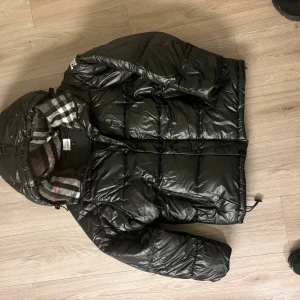 Burberry - Burberry Pufferjacka Snygg och elegant svart pufferjacka från Burberry, perfekt för kyliga vinterdagar. Jackan har en glänsande, quiltad yta och en klassisk huva fodrad med Burberrys ikoniska rutmönster. 