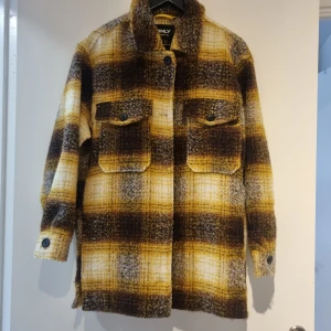 Rutig jacka från ONLY - Säljer en superfin rutig jacka från ONLY i storlek S. Den har en härlig mix av bruna, gula och vita nyanser. Jackan är långärmad med knappar framtill och två stora fickor på bröstet. Perfekt för höst och vår! Oanvänd men prislapp finns ej kvar. 