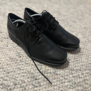 Svarta skinnskor från Rockport - Säljer ett par stilrena svarta skinnskor från Rockport i herrstorlek 44. Skorna har klassisk snörning och en bekväm sula med bra grepp. Perfekta för både vardag och festliga tillfällen. De är i mycket bra skick och redo för nya äventyr!