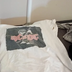 Vit AC/DC t-shirt - Säljer en vit AC/DC t-shirt med tryck av bandets logga och texten 'Shot in the Dark'. Perfekt för rockälskare! T-shirten är kortärmad och i bomull, vilket gör den bekväm att bära till vardags eller på konsert.