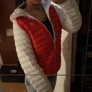 Röd och vit dunjacka från Haglöfs - Säljer en snygg och varm dunjacka från Haglöfs i rött och vitt. Jackan har en dragkedja framtill och en praktisk huva. Perfekt för kyliga höst- och vinterdagar. Den är i mycket bra skick och passar perfekt för utomhusaktiviteter.