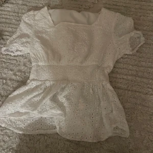 Vit blus med spetsdetaljer - Säljer en söt vit blus med spetsdetaljer och korta ärmar. Den har en elastisk midja som ger en fin passform och ett luftigt tyg med hålmönster. Perfekt för sommarens varma dagar eller en avslappnad vårlook. Passar både till jeans och kjol!