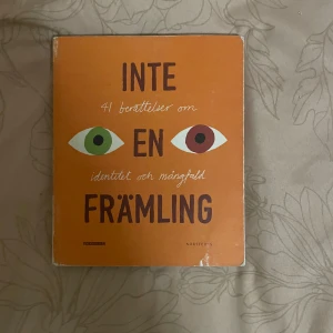 Inte en främling - Boken 'Inte en främling' innehåller 41 berättelser om identitet och mångfald. Den har en orange framsida med två ögon i olika färger. Boken utforskar teman som rättigheter, tillhörighet och rasism. Författare som Sofia Jannok och Rikard Wolff bidrar med sina perspektiv.