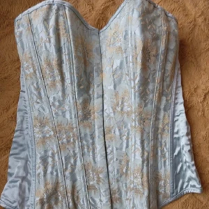 Elegant ljusblå korsett - Säljer en superfin ljusblå korsett med vackra broderade blommor i guld. Den har en klassisk passform och är perfekt för festliga tillfällen. Materialet känns lyxigt och ger en elegant look. Passar perfekt till både kjol och byxor!