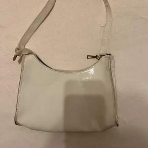 Vit axelväska med dragkedja - Säljer en stilren vit axelväska i syntetmaterial. Väskan har en praktisk dragkedja upptill och en enkel design som passar till många olika outfits. Perfekt för både vardag och fest! Säljs för 40