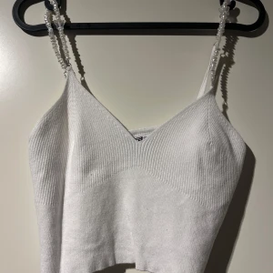 Vit ribbad linnetopp med pärlband - Säljer en superfin vit ribbad linnetopp med unika pärlband. Toppen är croppad och perfekt för sommardagar eller festliga tillfällen. Den har en stilren design med en v-ringad framdel och ärmlös stil. Passar perfekt till både jeans och kjol!