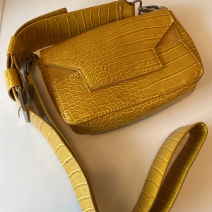 Handväska från Carin Wester med krokodilpräglat mönster - Handväska från Carin Wester. Säljer en superfin gul axelväska med ett coolt krokodilpräglat mönster. Väskan har en justerbar axelrem och en praktisk liten ficka framtill. Välanvänd men ändå fräsch. 