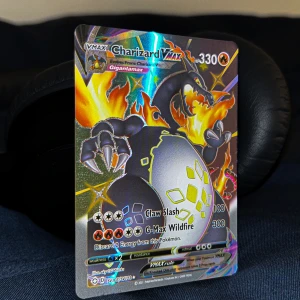 Charizard VMAX Pokémon-kort - Säljer ett Charizard VMAX Pokémon-kort med Gigantamax-design. Kortet har en cool och färgglad illustration av Charizard med eldflammor i bakgrunden. Det är i perfekt skick och har en holografisk yta som verkligen får det att sticka ut. Perfekt för samlare eller Pokémon-fans!