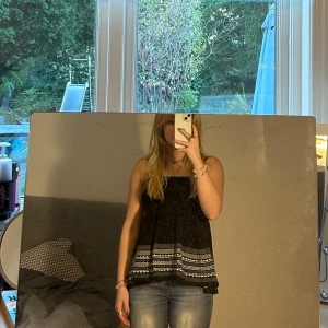 Svart topp från KappAhl - Säljer en svart topp från KappAhl med ett snyggt mönster i vitt och grått längst ner. Toppen är ärmlös och är taitare upptil och går ut nedtill