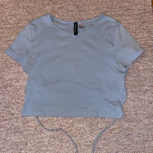 Blå bagtröja med knyt i ryggen. - Säljer en ljusblå T-shirt/ magtröja med knyt i ryggen. Använd nån gång men är i bra skick. Pris kan diskuteras.