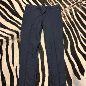 Mörkblå byxor från J.Lindeberg - Säljer ett par snygga mörkblå golfbyxor från J.Lindeberg. De har en tight fit och är även perfekta för både vardag. Byxorna har en knapp och dragkedja framtill och är i ett stilrent utförande.