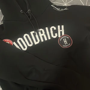 Svart hoodie från Hoodrich - Säljer en svart hoodie från Hoodrich med vit text på bröstet och en rund logga på sidan. Hoodien har en justerbar huva med snören och långa ärmar. Perfekt för en avslappnad stil. (passar strl S om man vill ha den lite mer lös)