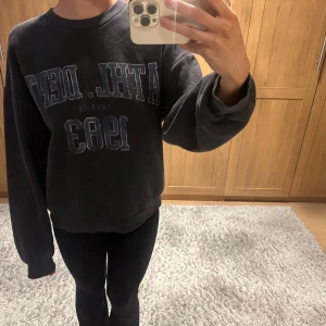 Svart sweatshirt med tryck - Säljer en svart sweatshirt med tryck i blått och vitt. Tröjan har långa ärmar och en avslappnad passform. Perfekt för en casual look.