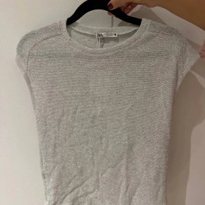 Vit kortärmad topp från Zara - Säljer en stilren silvrig topp från Zara i en blandning av polyester och bomull. Toppen har korta ärmar och en enkel design som passar perfekt till olika tillfällen. Ny med prislapp kvar.