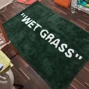 Grön matta med texten 'WET GRASS' - Snygg grön matta med vit text 'WET GRASS' i stora bokstäver. Perfekt för att ge rummet en unik och modern touch. Passar bra i vardagsrummet eller sovrummet för en stilren look. Width 133x Length 195m