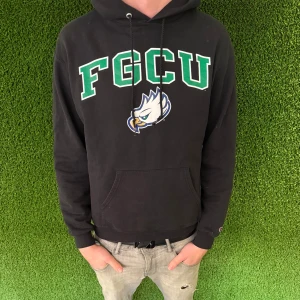 Svart college hoddie - Snygg svart hoodie med FGCU-logga i grönt och vitt framtill. Tröjan har en klassisk känguruficka och justerbar huva med dragsko. Perfekt för en avslappnad stil.