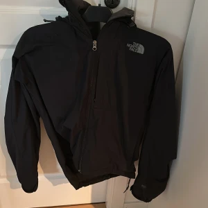 Svart vindjacka från The North Face - Säljer en svart vindjacka från The North Face med huva och dragkedja. Jackan har en stilren design och är perfekt för blåsiga dagar. Den har långa ärmar och är tillverkad i ett lätt material som skyddar mot vind. Drags kedjan är lite trög nere på jackan men annars i väldigt bra skick
