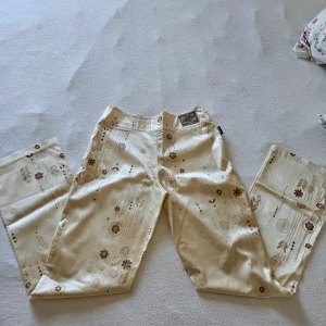 Beige byxor med blommönster från Amnesia original Paris - Snygga beige byxor från Amnesia original Paris med ett unikt blommönster i guld, vitt och brunt. Byxorna har en rak passform och är dekorerade med knappar i midjan. Perfekta för en stilren look. Tillverkad i acetat som är konstgjord silke - vekfri,  andningsbar och lättskött material! Stl 42 fransk, som betyder att de passar till stl 38/40