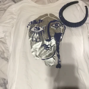 Vit t-shirt med ansiktsmotiv från Zara - Säljer en vit t-shirt från Zara med ett unikt ansiktsmotiv i silver och svart. T-shirten har korta ärmar och är perfekt för en trendig look. Passar bra till både jeans och kjol.