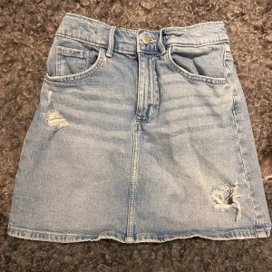 Blå jeanskjol från H&M - Säljer en cool blå jeanskjol från H&M med slitna detaljer och klassisk femficksdesign. Kjolen har en knapp och dragkedja framtill. strl 140 men passar Xxs