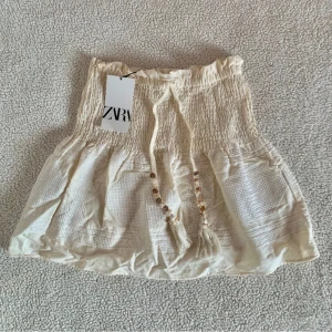 Vit kjol med volangdetaljer - Söt vit kjol med volangdetaljer och knytband i midjan. Perfekt för varma sommardagar. Den har en elastisk midja för bekväm passform.