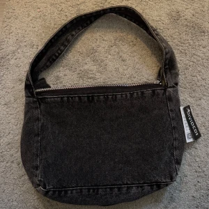 Svart axelväska från Divided - Snygg svart axelväska i jeansmaterial från Divided. Väskan har en praktisk dragkedja upptill och en rymlig insida med extra fack. Perfekt för att bära med sig det nödvändigaste med stil.