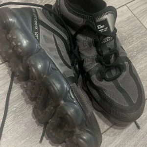 Grå Nike Vapormax sneakers - Snygga grå Nike Vapormax sneakers med svart detaljering och transparent sula. Skorna har snörning och en modern design som passar perfekt för en sportig look.