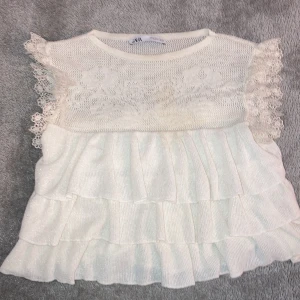 Vit spetsblus från Zara - Säljer en söt vit blus från Zara med spetsdetaljer och volanger. Blusen har korta ärmar med spets och flera lager av volanger som ger en romantisk look. Perfekt för en somrig stil!