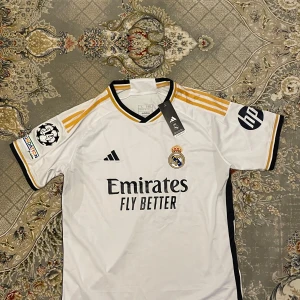 Real Madrid Fotbollströja  - Säljer en vit Real Madrid fotbollströja från Adidas med svarta och gula detaljer. Tröjan har korta ärmar och texten 'Emirates Fly Better' på framsidan. Baksidan har namnet 'Bellingham' och nummer 5. Perfekt för fotbollsfans!