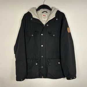 Fjällräven jacka - Säljer en svart jacka från Fjällräven med huva och mjukt foder. Jackan har knappar och dragkedja framtill samt två bröstfickor med knappar. Perfekt för kyliga dagar. Storlek XS men passar som S. Skriv vid ytterligare frågor eller funderingar🤝