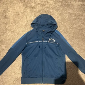 Blå huvtröja från BOSS - Snygg blå huvtröja från BOSS med dragkedja och broderad logga på bröstet. Tröjan har långa ärmar och en praktisk huva. Perfekt för en avslappnad stil.