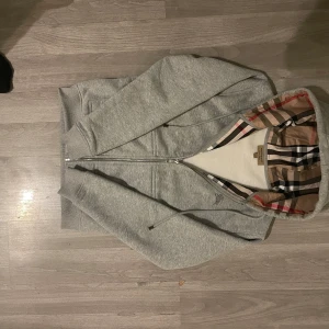Grå huvtröja från Burberry - Säljer en stilren grå huvtröja från Burberry med dragkedja och klassiskt rutigt mönster i huvan. Tröjan har en broderad logga på bröstet och ribbade muddar vid ärmslut och nederkant. Perfekt för en avslappnad look. Priset kan såklart diskuteras