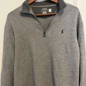 Grå tröja från Polo Ralph Lauren - Säljer en stilren grå tröja från Polo Ralph Lauren med dragkedja vid halsen. Tröjan har en broderad logotyp på bröstet och är perfekt för en avslappnad look. Är knappt använd och är som helt ny 