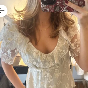 Vit blommig blus - Charmig vit blus med blommigt mönster och volangdetaljer. Blusen har korta ärmar och en romantisk offshoulder-design, perfekt nu för våren och sommaren. 