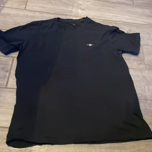 Svart t-shirt från GANT - Säljer en stilren svart t-shirt från GANT med ett litet tryck av deras logotyp på bröstet. Perfekt för en avslappnad look.