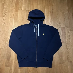 Mörkblå zip hoodie från Polo Ralph Lauren - Säljer en snygg mörkblå zip hoodie från Polo Ralph Lauren med dragkedja och det klassiska gula logotypen på bröstet. Tröjan har långa ärmar och en justerbar huva med vita snören. En pytteliten nästan osynlig färgfläck på luvan. Perfekt för en avslappnad stil.