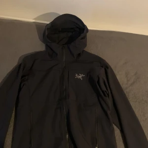 Arc'teryx jacka - Säljer en stilren svart jacka från Arc'teryx med dragkedja och huva. Jackan har en broderad logotyp på bröstet och är perfekt för utomhusaktiviteter. Den är långärmad och har en modern design.
