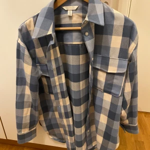 Blå och vit rutig skjorta från H&M - Säljer en stilren blå och vit rutig skjorta från H&M. Skjortan har långa ärmar och knappar framtill. Perfekt för en avslappnad look. Den är stor i storleken 