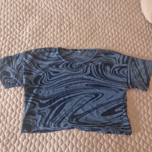 Blå mönstrad croppad topp - Säljer en blå croppad topp med ett unikt abstrakt mönster i mörkare nyanser. Toppen har korta ärmar och en rund halsringning, perfekt för en avslappnad stil.