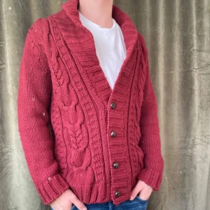 Röd stickad kofta med knappar - Säljer en mysig röd stickad kofta med knappar framtill. Koftan har ett snyggt flätmönster och ribbade muddar vid ärmslut och nederkant. Perfekt för kyliga dagar!