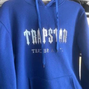 Trapstar hoodie  - En blå huvtröja med en silverfärgad logotyp på framsidan. Huvtröjan har en huva med dragsko och en ficka på magen.