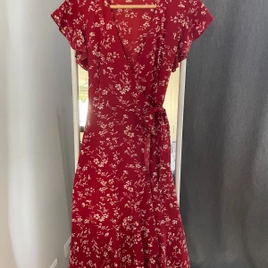 Röd blommig klänning från Shein - Säljer en superfin röd klänning från Shein med ett vackert blommigt mönster. Klänningen har en omlott-design med knytband i midjan och korta ärmar. Perfekt för sommarens alla fester eller en mysig vårdag. Den är i storlek XS och sitter riktigt bra!
