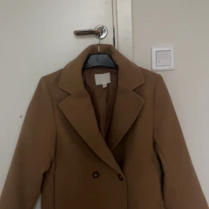 Beige kappa - Snygg beige kappa med klassisk krage och dubbelknäppning. Perfekt för höst och vår, med en stilren design som passar till många tillfällen. Jag köpte den förra året och har använt den typ fyra gånger. Den är ifrån HM. Används inte därför säljer jag den. Den har alla knappar och det finns inga hål och den är ganska tjock. 