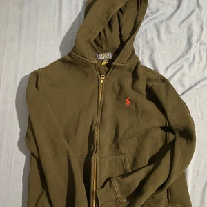 Polo Ralph Lauren zip hoodie 11-14 år - Säljer en olivgrön hoodie från Polo Ralph Lauren med dragkedja och den klassiska röda loggan på bröstet. Perfekt för en avslappnad stil med en praktisk huva och fickor framtill.