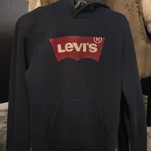 Mörkblå hoodie från Levi's - Säljer en snygg mörkblå hoodie från Levi's med det klassiska röda och vita logotyptrycket på bröstet. Tröjan har en praktisk magficka och en bekväm huva. Perfekt för en avslappnad stil.