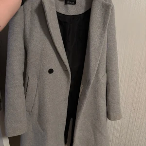 Grå kappa från Bershka - Snygg grå kappa från Bershka med klassisk design och knappar framtill. Perfekt för höst och vår. Kappan har långa ärmar och två fickor på sidorna. Sprucken på insidan men inget som påverkar funktionen