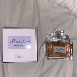 Miss Dior Eau de Parfum - Elegant och feminin parfym från Dior. Flaskan har en stilren design med en silverfärgad rosett och en ljusrosa vätska. Förpackningen är vit med lila detaljer och den ikoniska Dior-loggan.