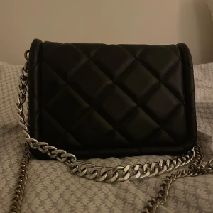 Svart quiltad axelväska med kedja - Snygg svart axelväska i quiltat mönster med en robust kedja som axelrem. Väskan har en praktisk storlek och stängs med magnetknappar. Perfekt för att ge en stilren touch till din outfit. Fint skick, enda defekten är att kedjan tappat lite färg!☺️